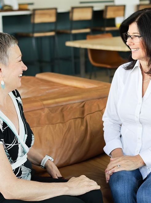Two women laughing, seated close together on a large leather cushion indoors, possibly an office.