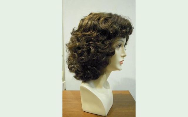 Parrucca su testa di manichino, capelli castano scuro e ricci. Manichino beige su un tavolo di legno.