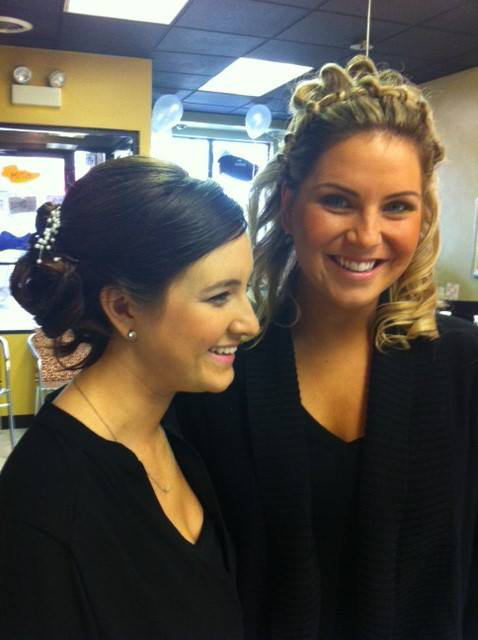 Two smiling women standing indoors, one with dark hair and one with blonde curls, in a brightly lit room