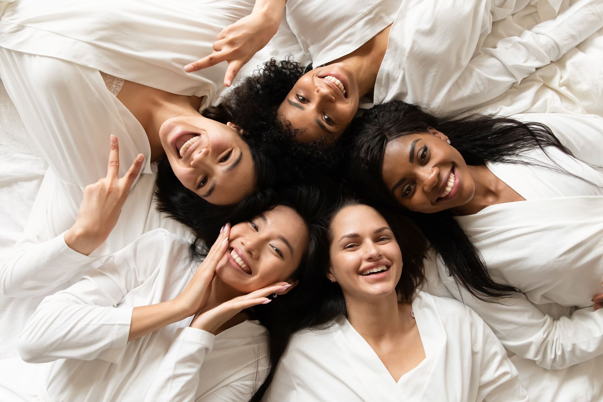 Five friends lying in a circle on white bedding, smiling and waving at the camera.