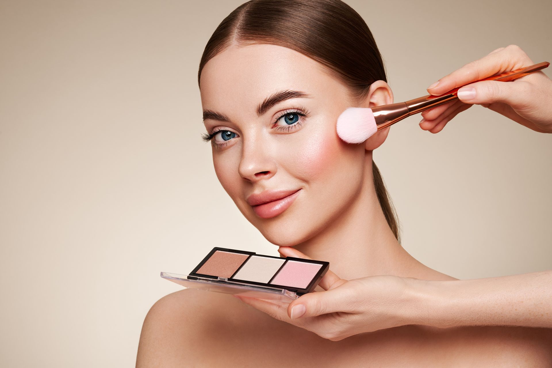 Woman applying pink blush with a makeup brush while holding a makeup palette on a neutral background