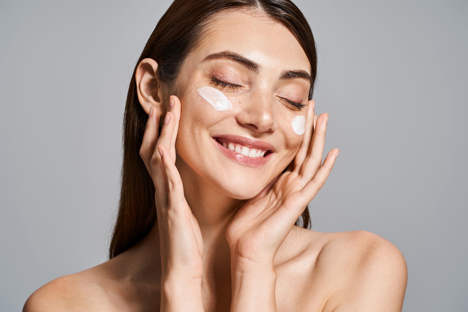 Smiling woman applying cream to her cheeks against a gray background