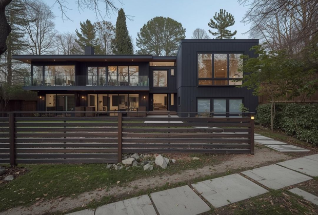 A modern, two-story, dark-sided house with expansive windows and a slatted wooden fence, set in a wooded area at dusk.