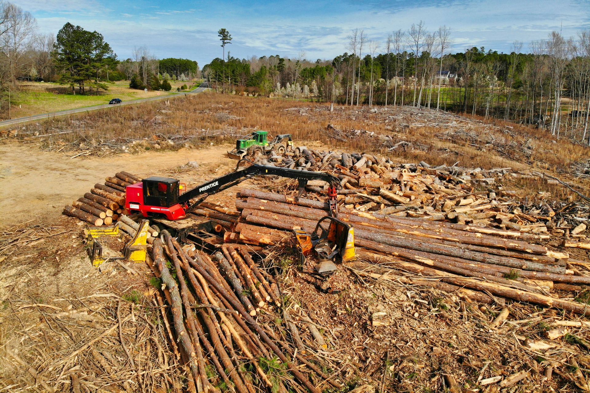 A red forestry machine with a grapple arm moves logs in a cleared, harvested woodland area under a blue sky.