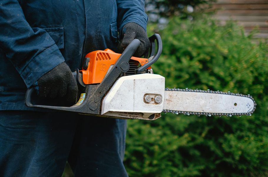 A person in dark work clothes and gloves holds an orange and white chainsaw in front of a green bush.