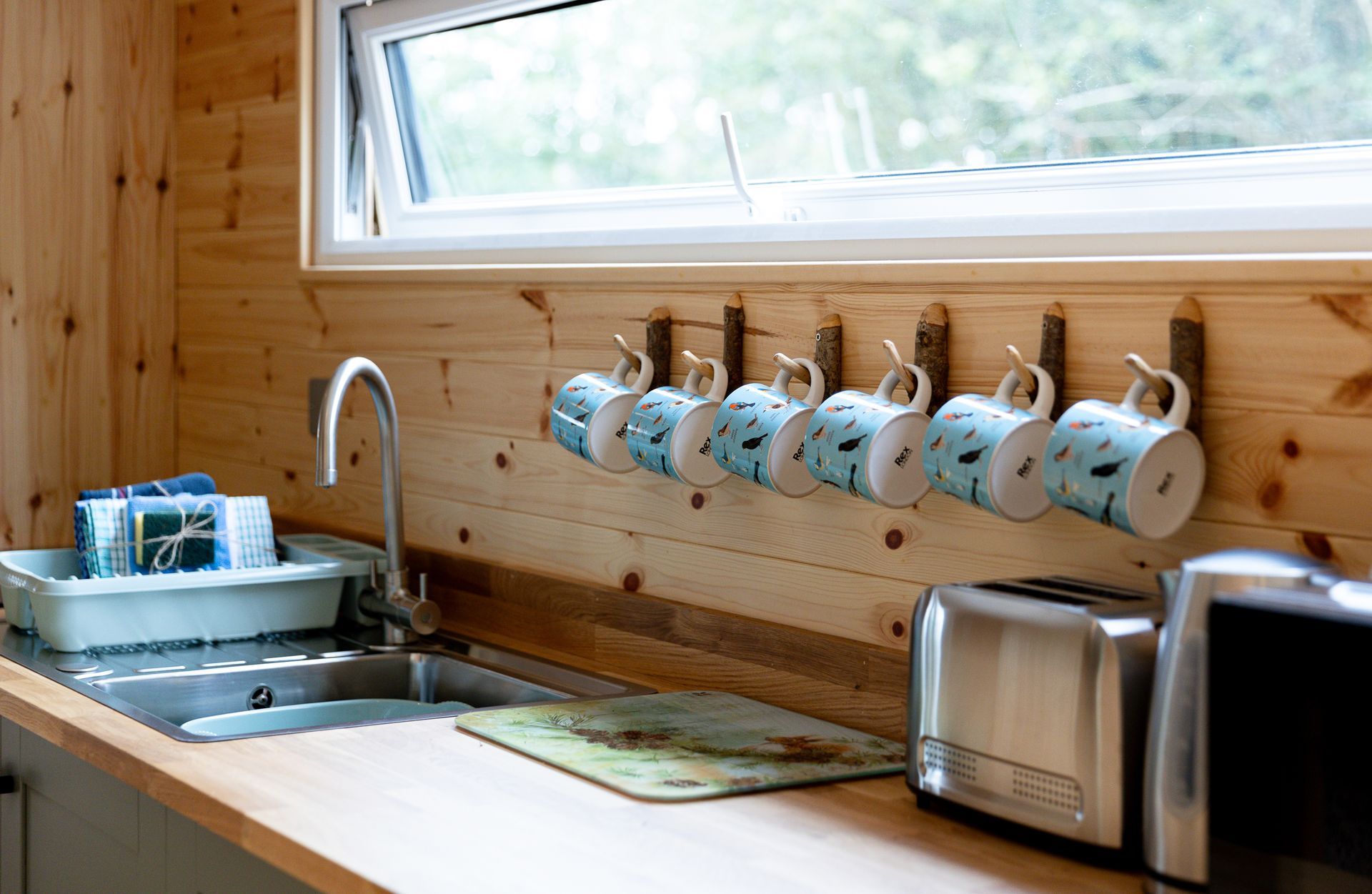 Kitchen interior with wooden walls, sink, mugs on hooks, and a toaster.