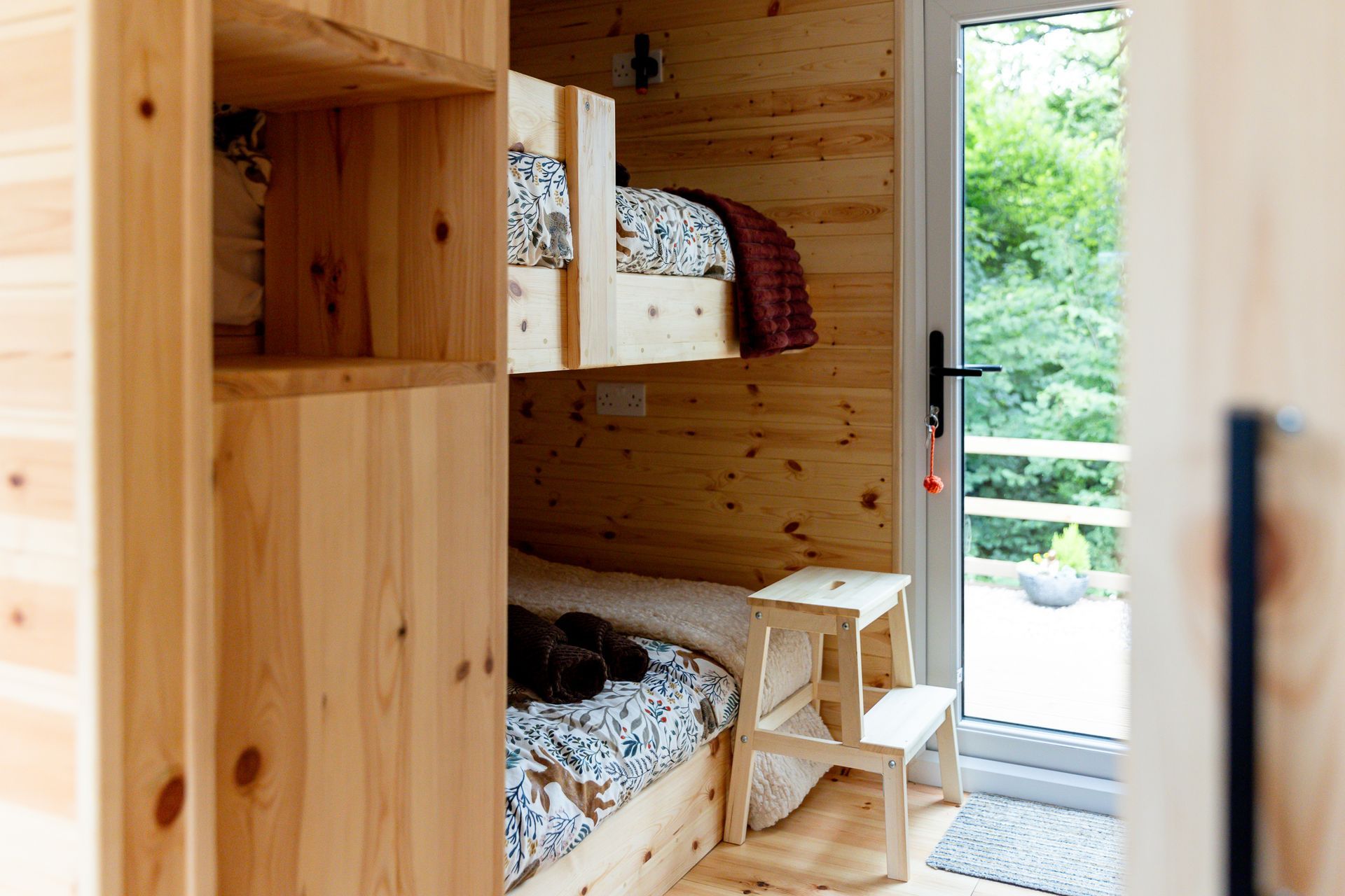 Wooden bunk beds in a cabin. Doorway to exterior view. Small wooden step stool. Natural wood tones.