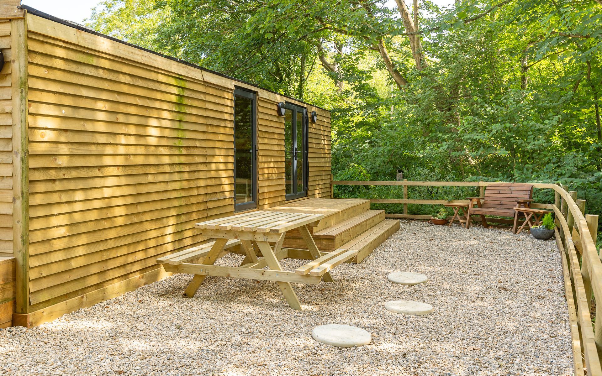 Wooden cabin with picnic table on a gravel patio, surrounded by a wooden fence and trees.