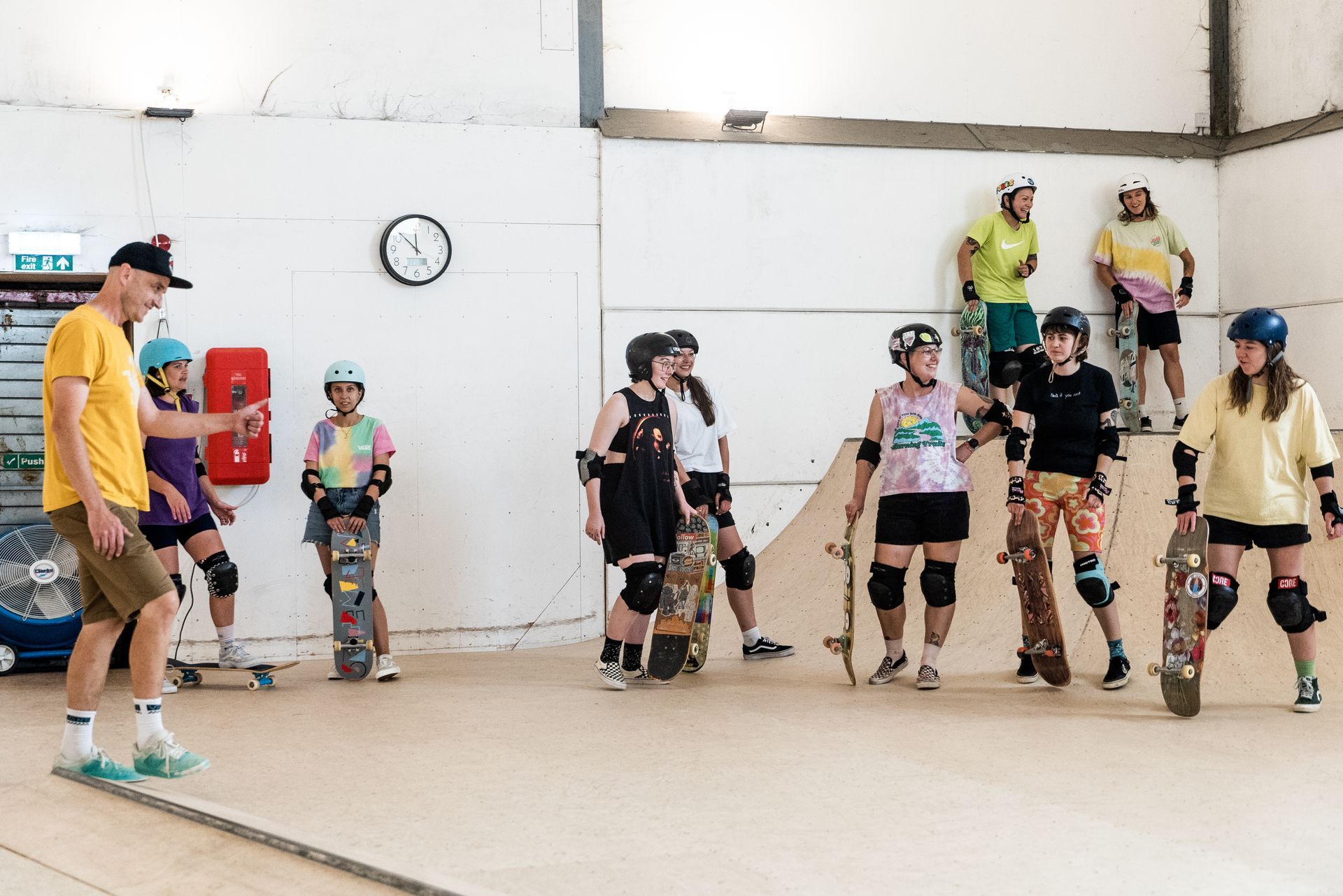 A group of people with skateboards in an indoor skatepark; a man is coaching.