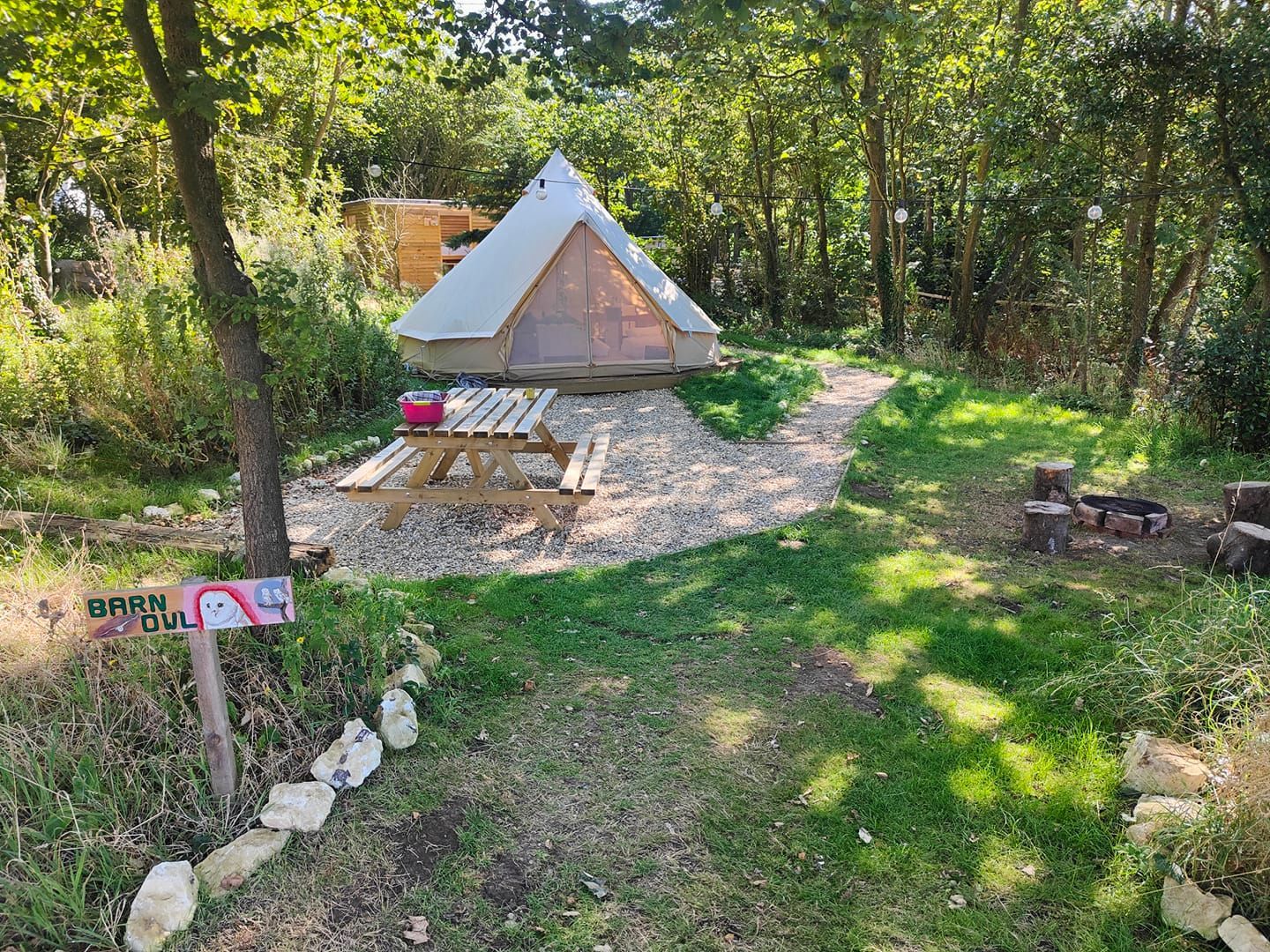 A campsite with a triangular tent, picnic table, and fire pit in a wooded area.