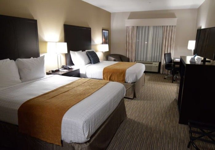 A hotel room featuring two queen-sized beds with white linens and tan runners, a dark wood dresser, desk, and lamp.