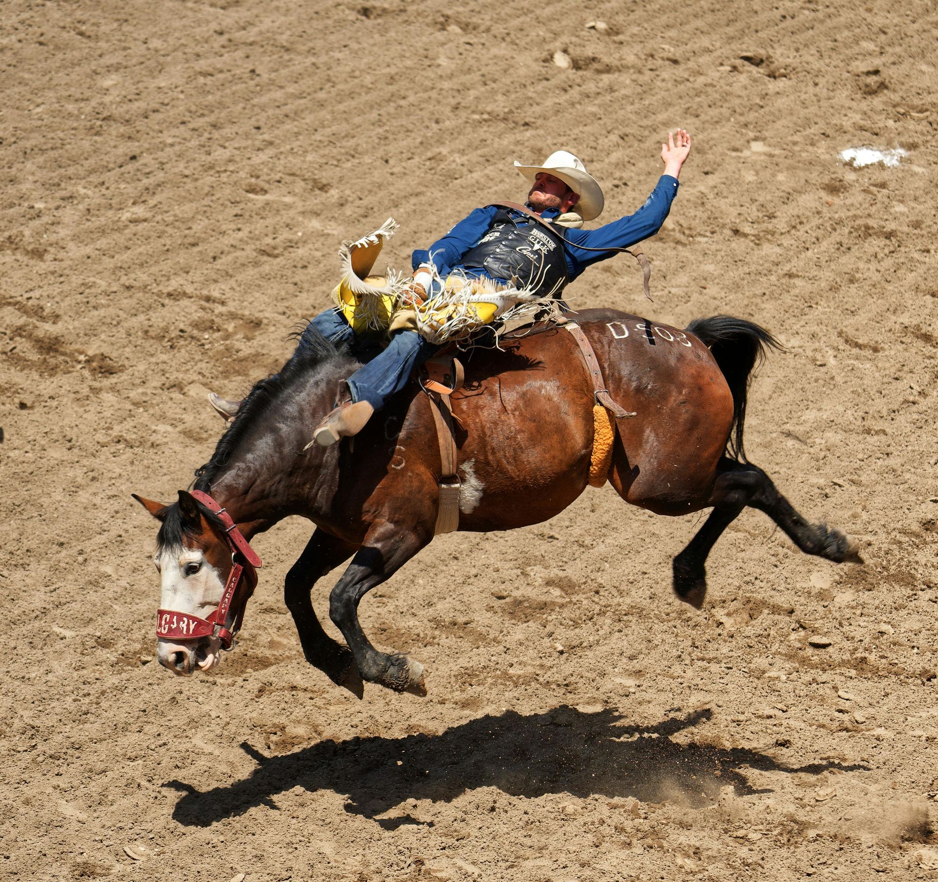 A cowboy riding a bucking brown horse in a dirt arena during a rodeo.