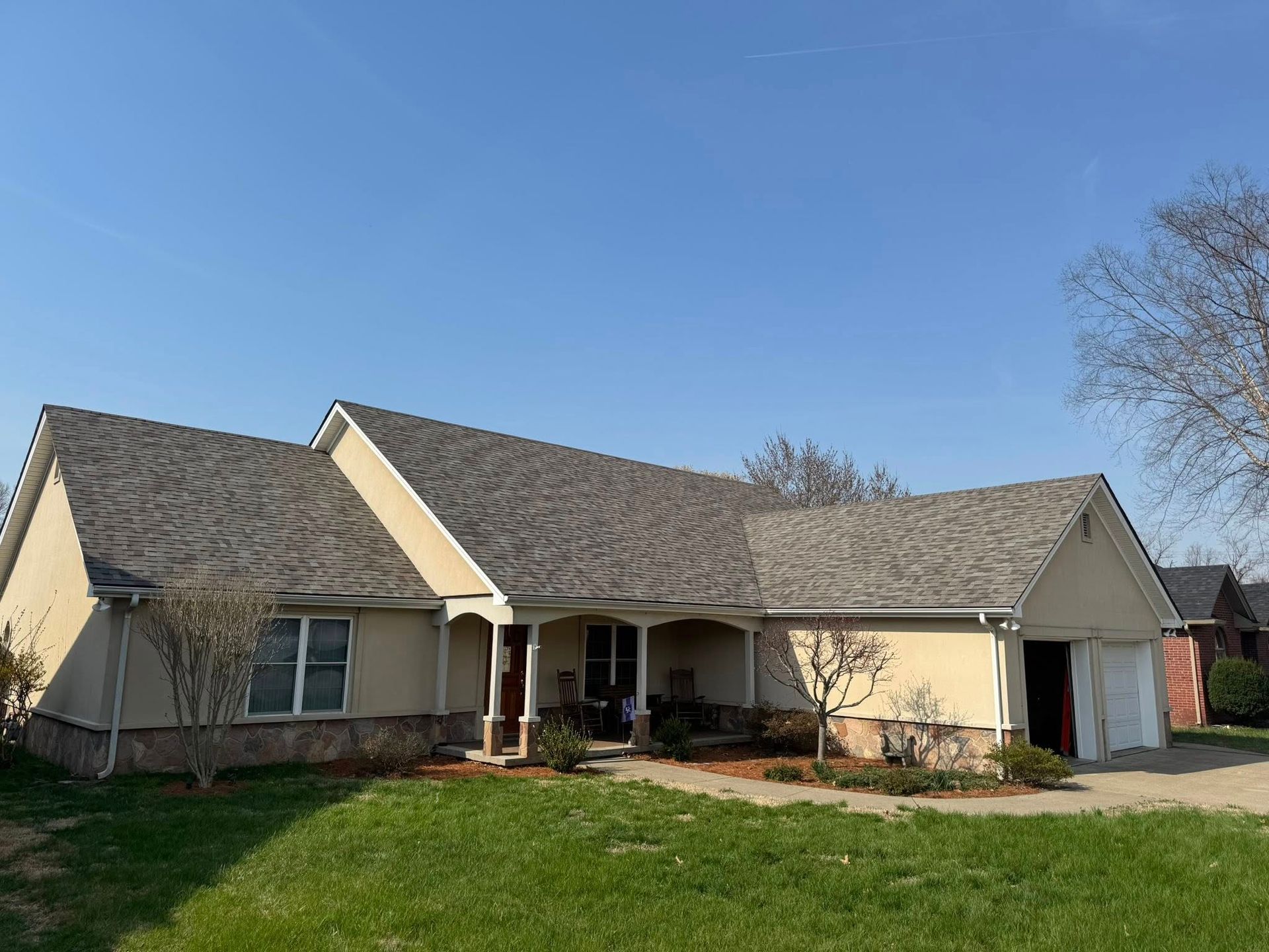 Single-story beige suburban house with a gray roof, front porch, and attached garage under a clear blue sky