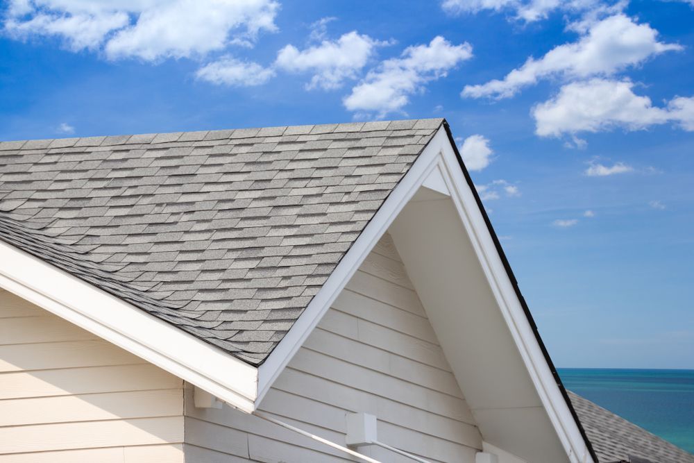 Gray shingled house roof with white trim beside a blue ocean under a partly cloudy sky