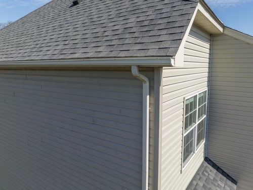Exterior corner of a gray house with vinyl siding, shingled roof, and a white gutter and window