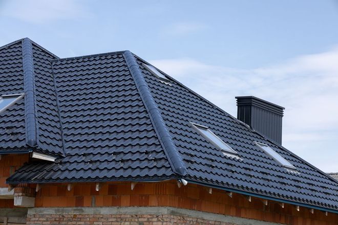 Dark blue tiled roof of a house with dormer windows and a brick chimney against a cloudy sky