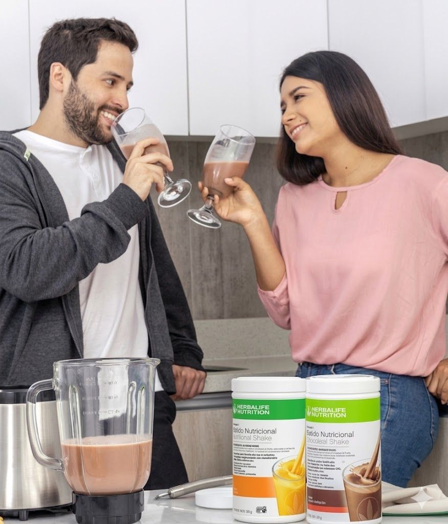 Dos personas sonriendo y brindando con batidos de proteínas en una cocina junto a recipientes de suplementos y una licuadora.