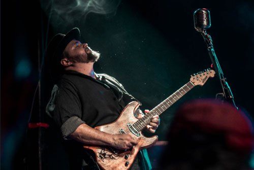 Image contains man playing guitar, hat, beard, smoke, carbon microphone