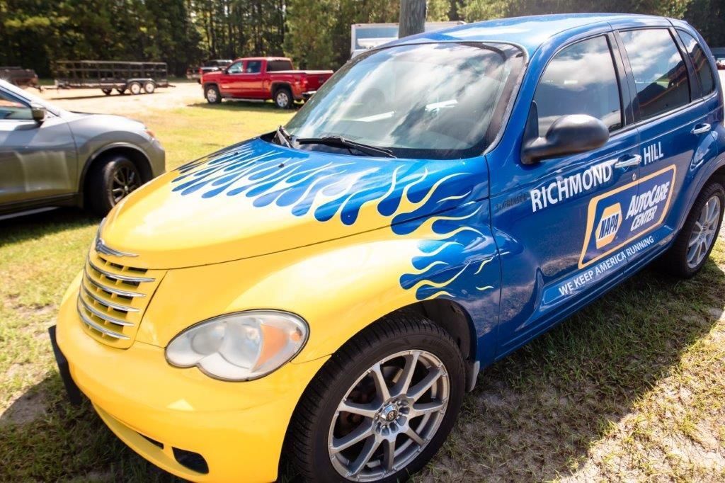 Blue and yellow promotional compact car parked on grass, decorated with wave graphics and logos.