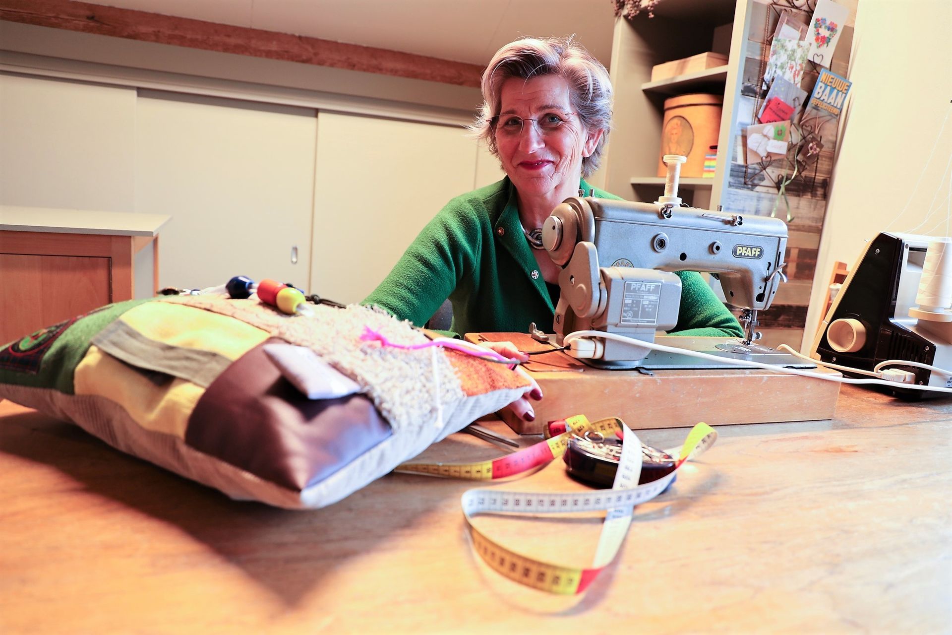Iemand zit te naaien aan een naaimachine in een gezellig knutselkamertje, met een speldenkussen en stof op tafel.