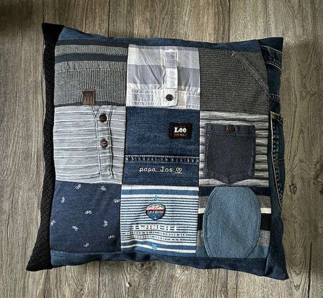 Een herinneringskussen van spijkerstof met patchwork in blauw, grijs en zwart op een houten vloer.