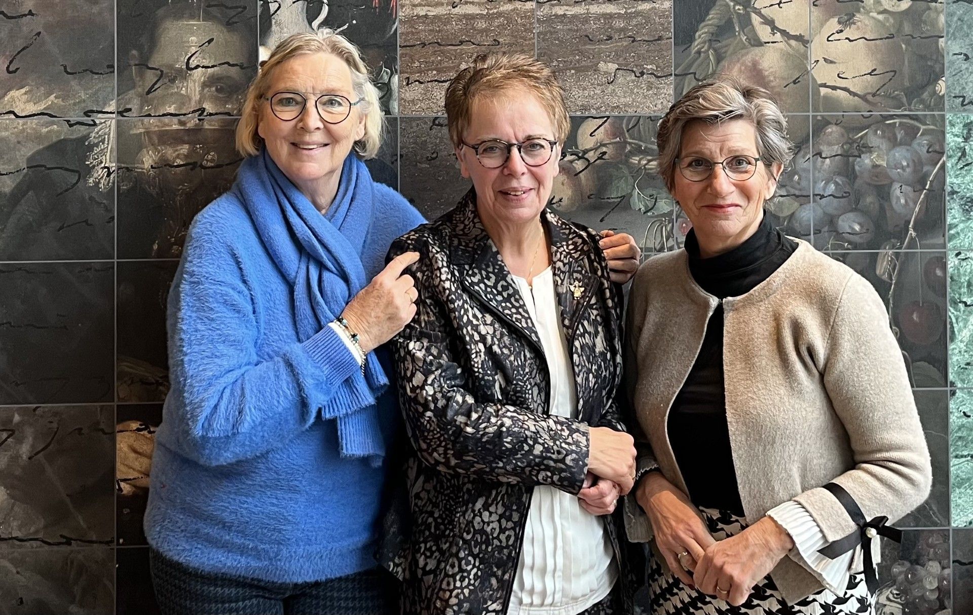 Drie vrouwen staan ​​dicht bij elkaar, glimlachend, tegen een stenen muur.