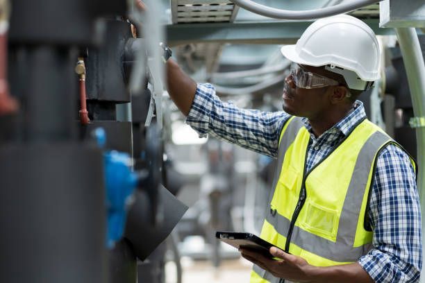A person in safety gear inspecting equipment with a tablet in an industrial setting.