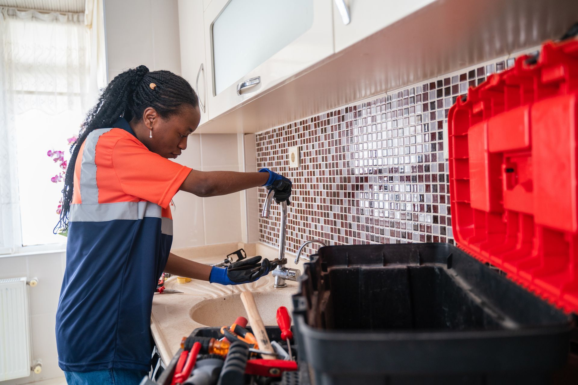 Woman in work clothes working on a kitchen faucet with tools from a toolbox.