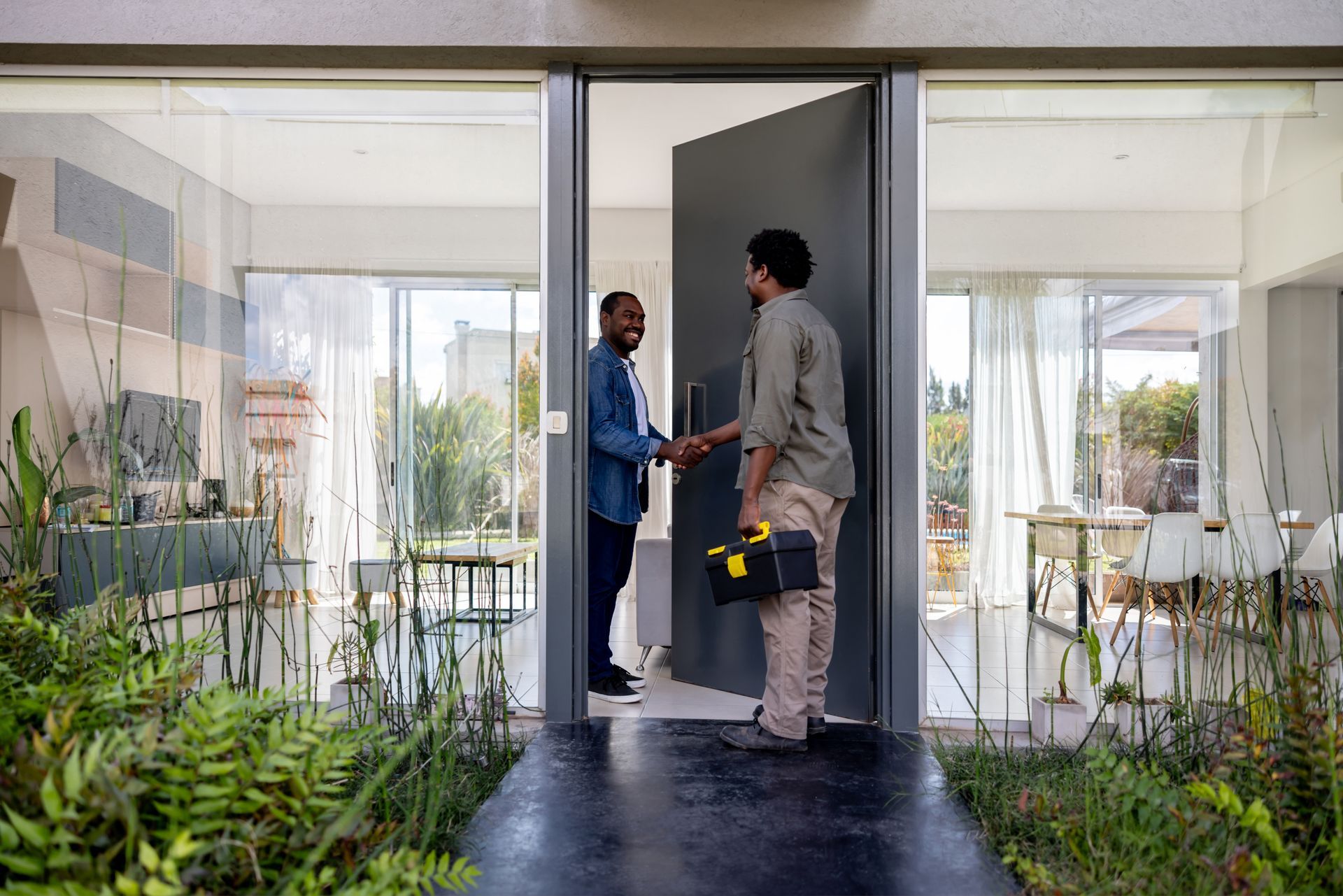 Man greets worker at front door, shaking hands. Worker holds a toolbox, inside a modern home.