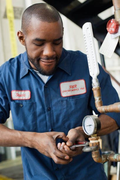 Man in blue work shirt, using a wrench on pipes with a gauge. Industrial setting.