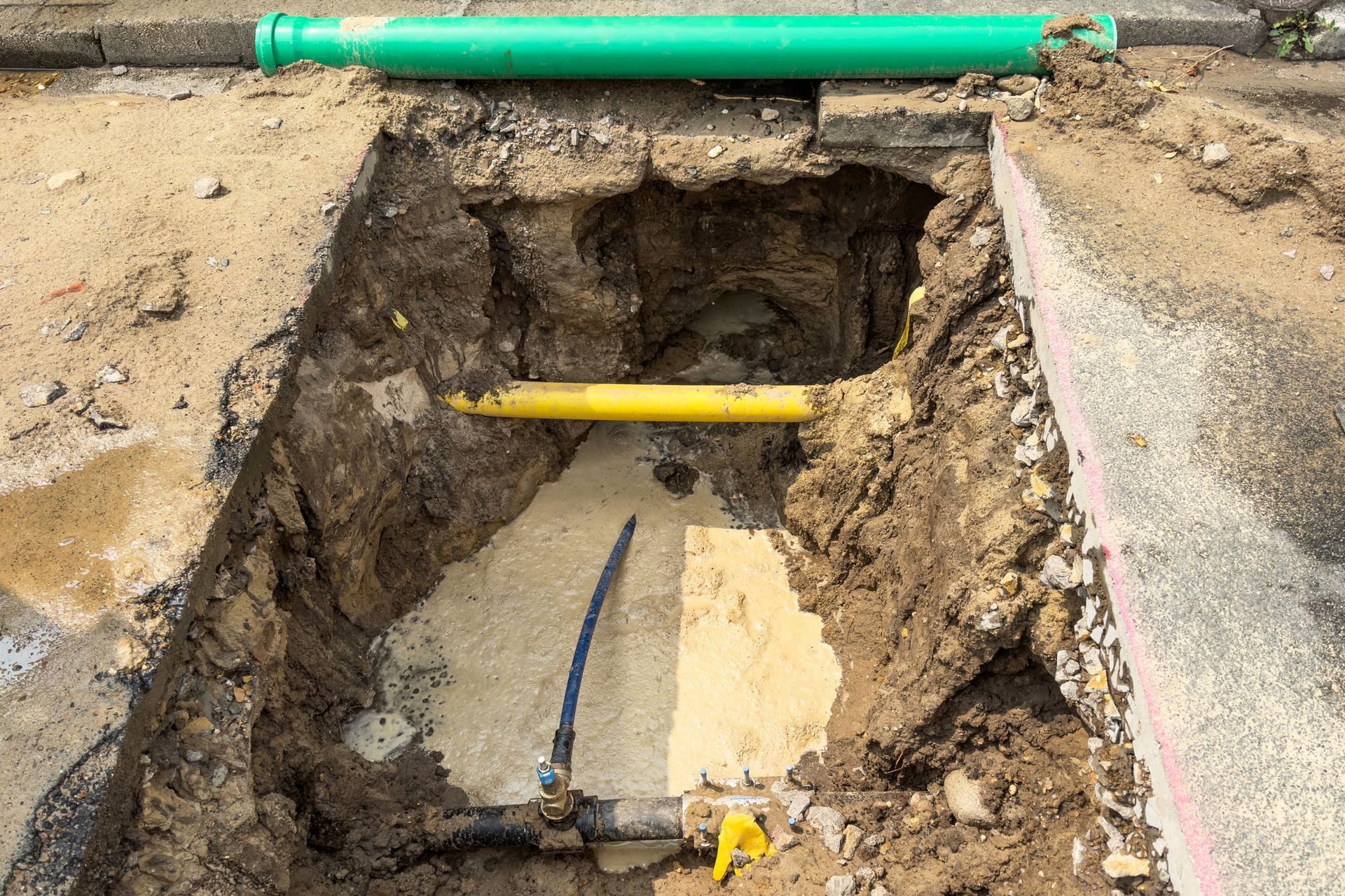 Open trench in asphalt with green and yellow pipes.