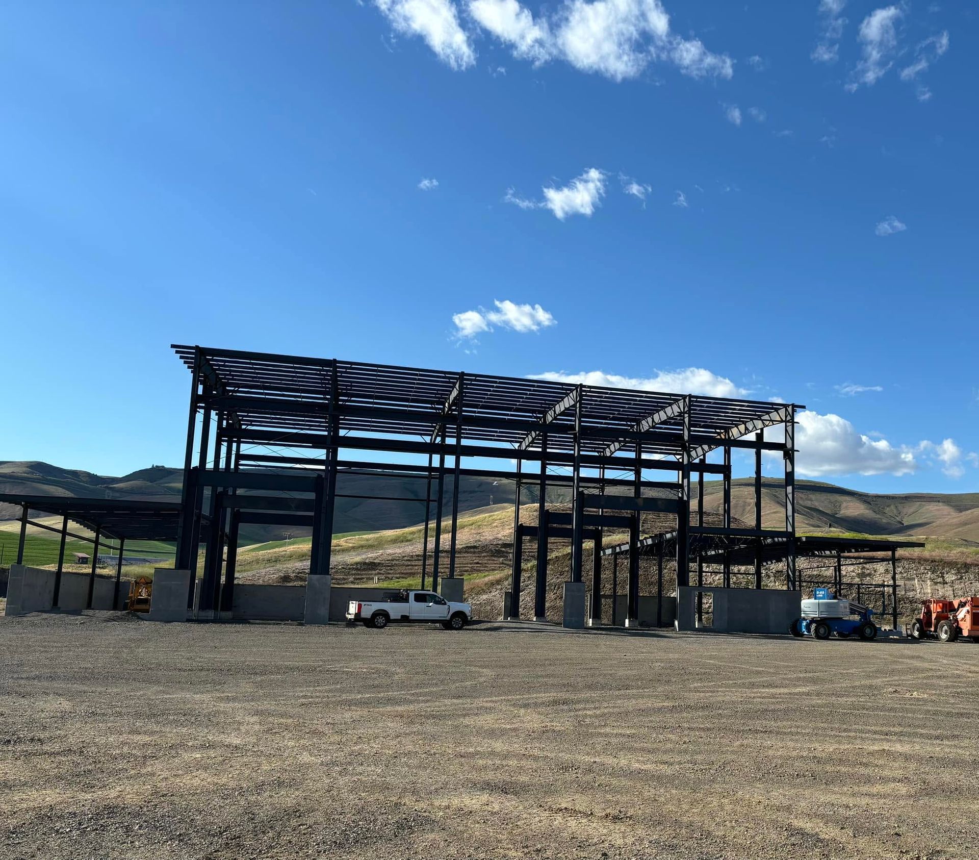 Steel-framed structure under construction in a gravel lot beneath a blue sky with scattered clouds.