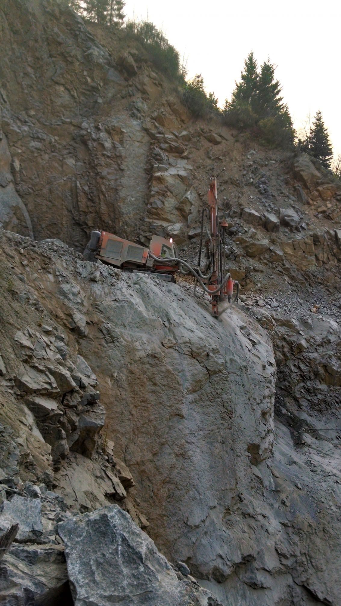 Rocky cliffside with a red drilling rig working on the slope near trees above.