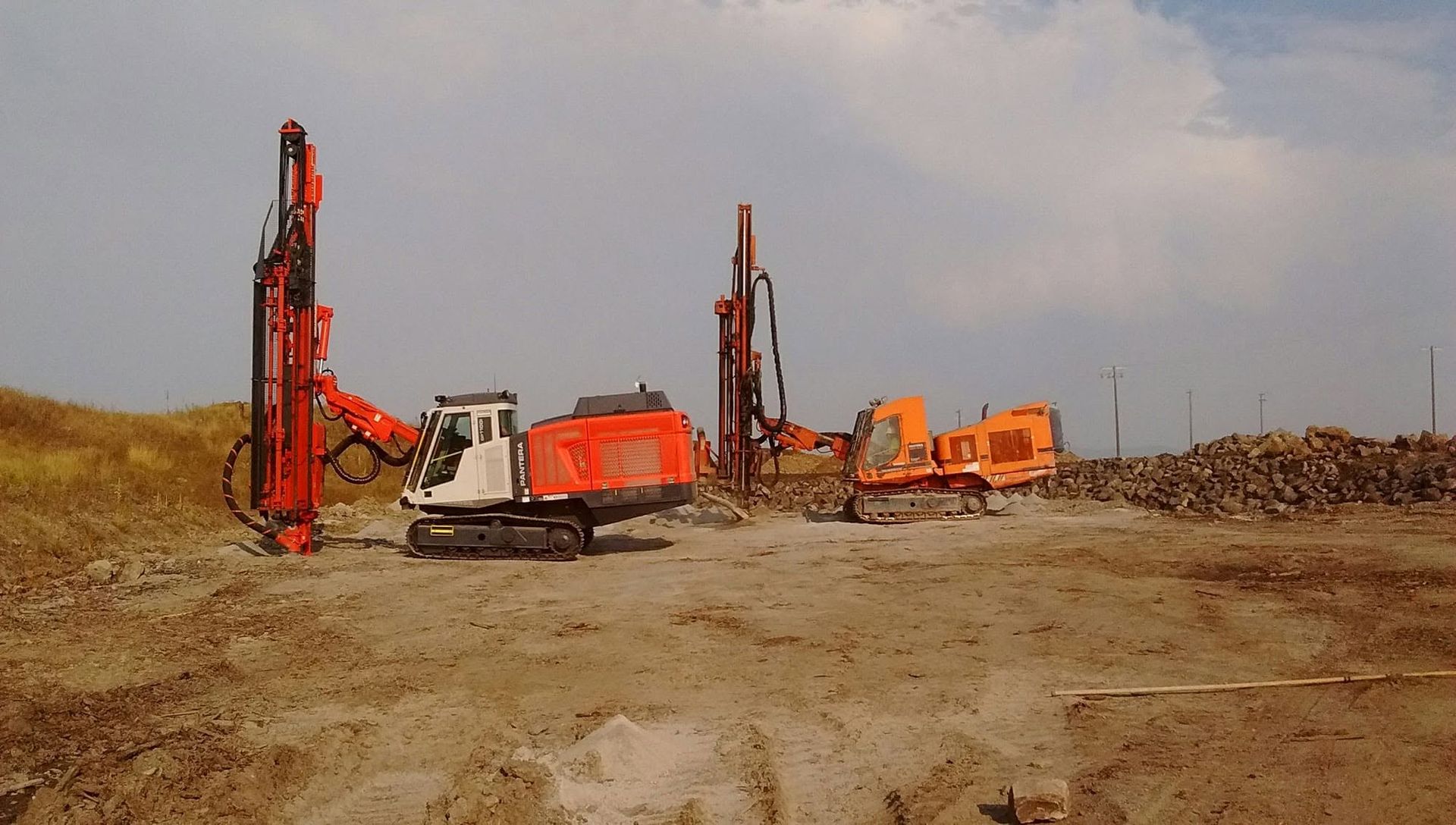 Orange drilling machines on a barren dirt construction site under a cloudy sky