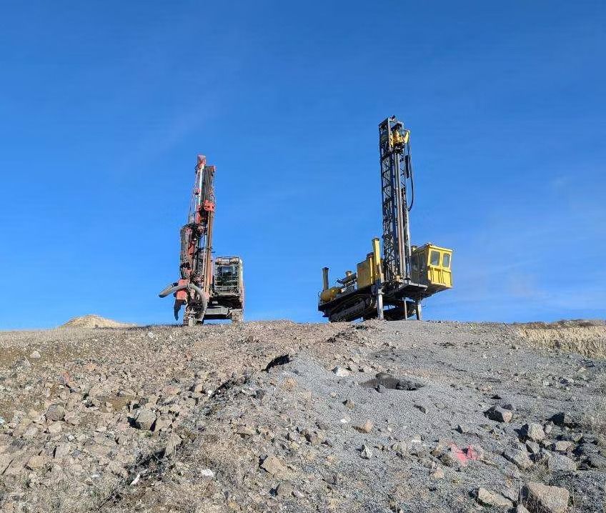 Two drilling rigs on a rocky construction site under a clear blue sky
