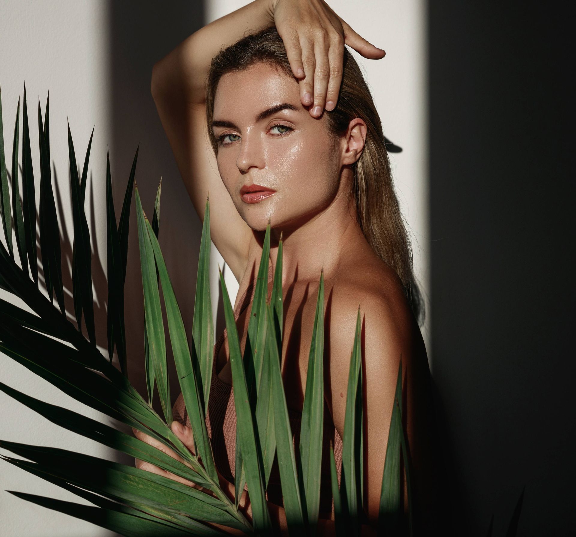 Portrait of a model with slicked-back hair, posing behind green palm leaves against a dark background