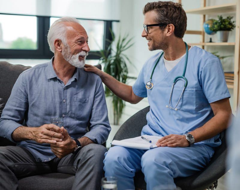 An elderly man is sitting on a couch talking to a doctor.
