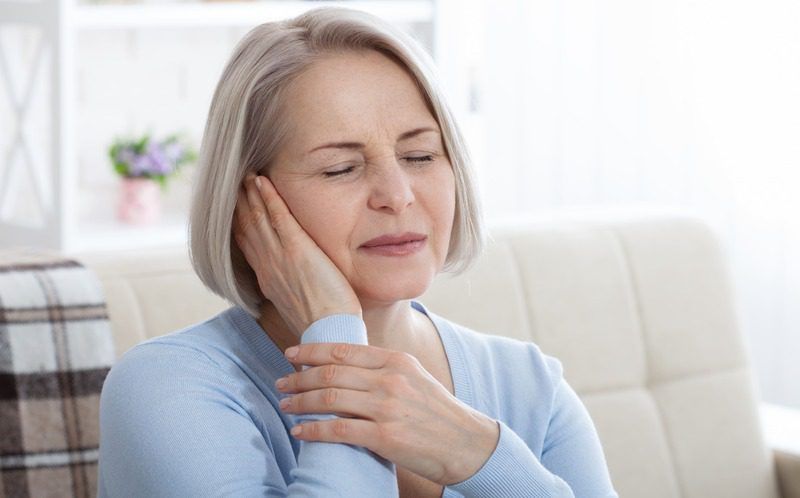 A woman is sitting on a couch holding her ear because she has a toothache.