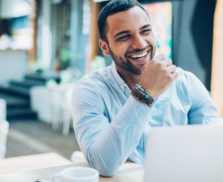 A man is sitting at a table with a laptop and smiling.