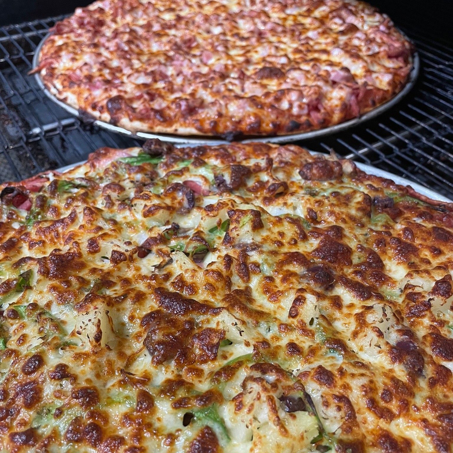 Two cheese pizzas baking on a grill, with browned, bubbly tops and one in the foreground.