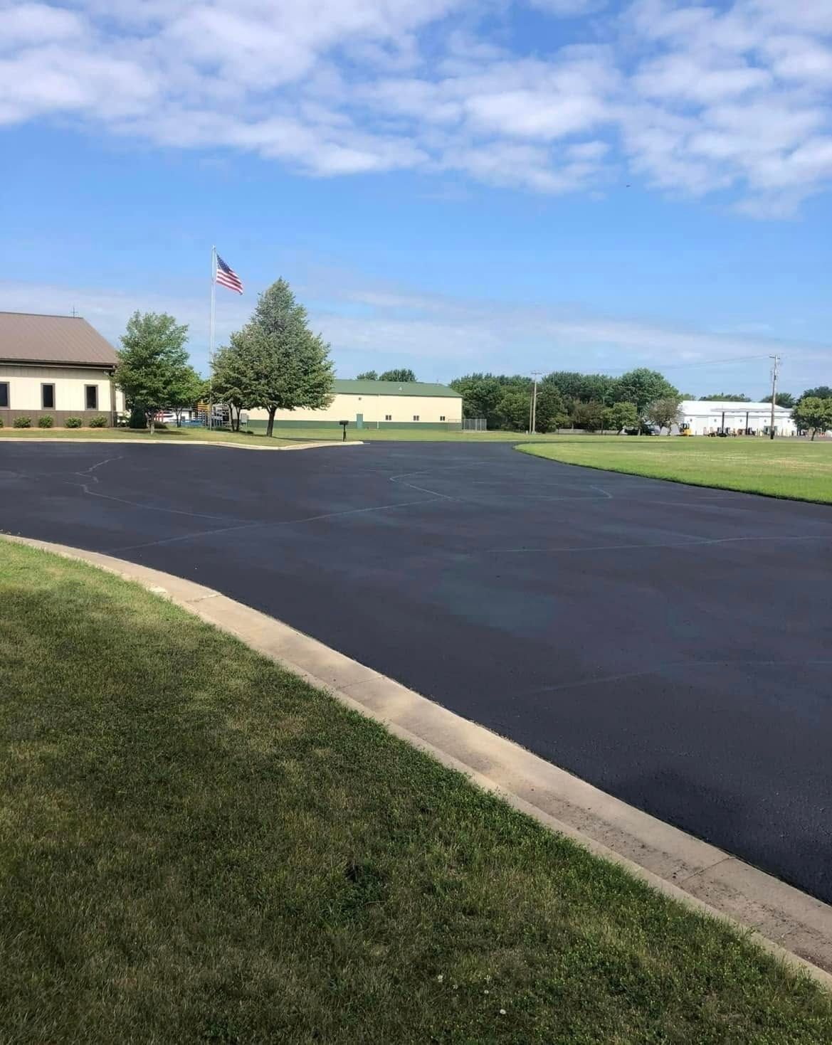 A freshly paved, dark asphalt parking lot bordered by a concrete curb and green grass under a blue sky with scattered clouds.