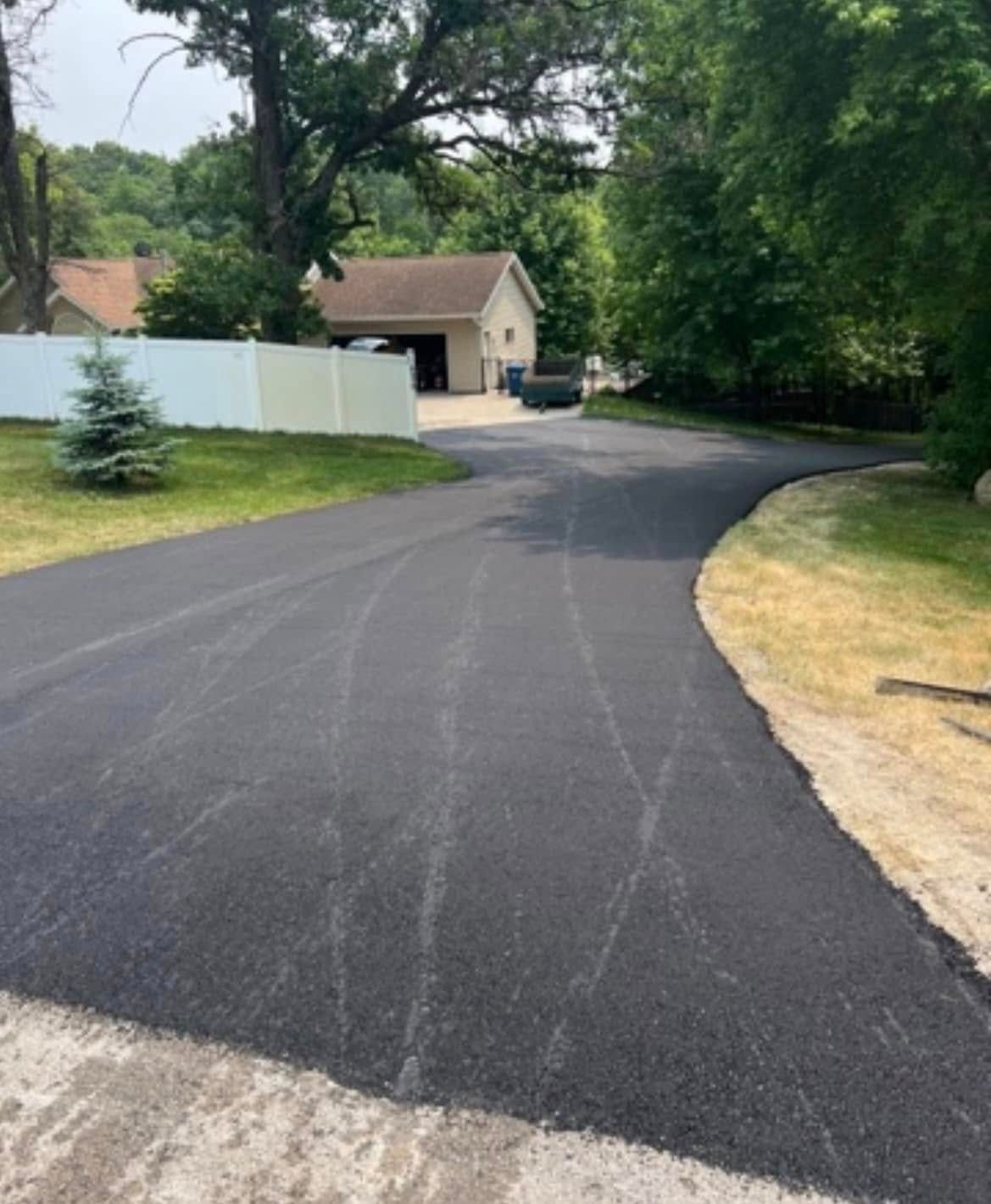 A freshly paved black asphalt driveway leads toward a house with a garage, flanked by a white fence and lush green trees.