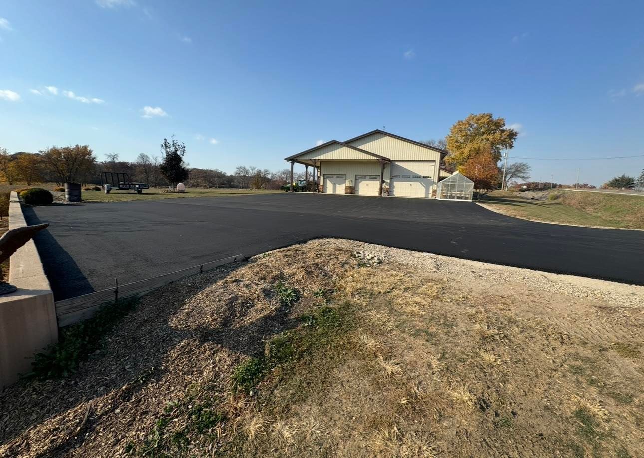 A wide, freshly paved asphalt driveway leads to a light-colored detached garage under a clear blue sky.