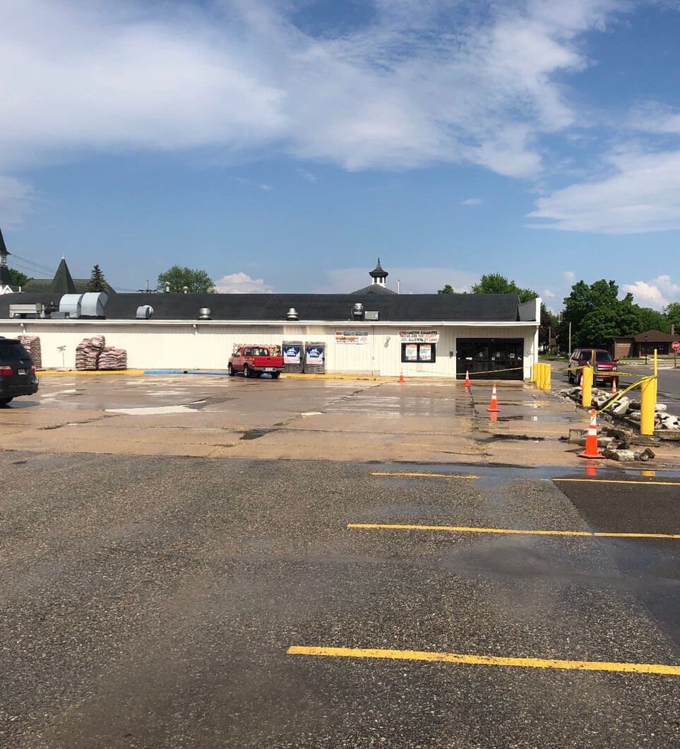 A white commercial building with a dark roof and a wet parking lot containing a red truck and orange traffic cones.