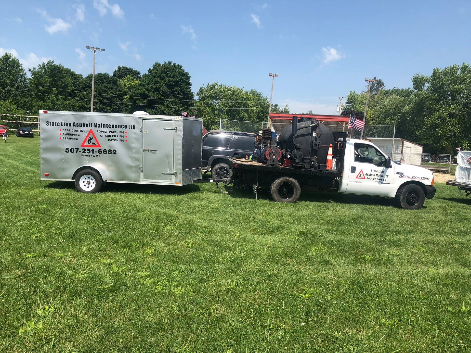 A white utility truck towing a silver trailer parked on a grassy field under a sunny sky.