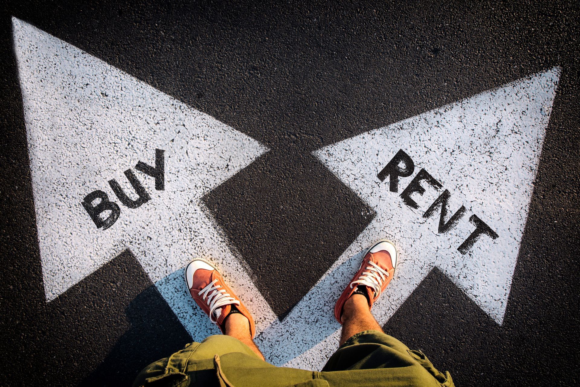 Feet at a fork in the road between “BUY” and “RENT” arrows on asphalt.