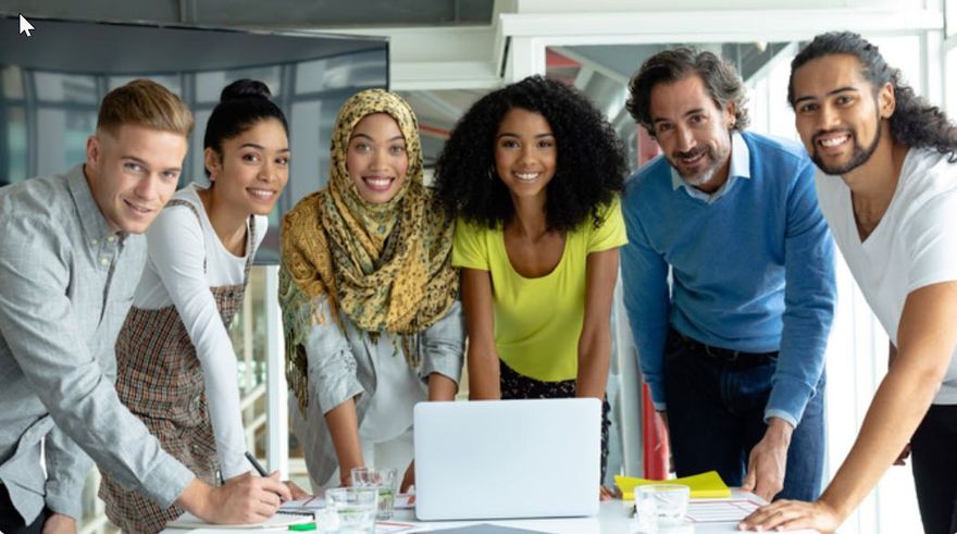 Group of six coworkers leaning over a laptop in a bright office, smiling at the camera.