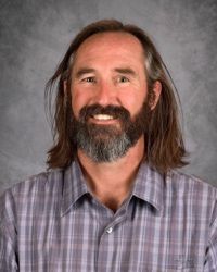 Smiling bearded man with shoulder-length hair in a plaid shirt against a gray backdrop