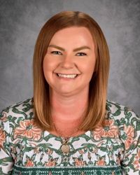 Smiling person with straight auburn hair wearing a green floral top against a gray backdrop