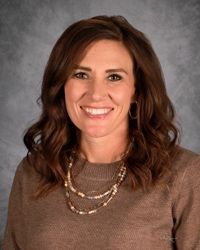 Smiling woman in a brown sweater and layered necklace against a gray studio backdrop